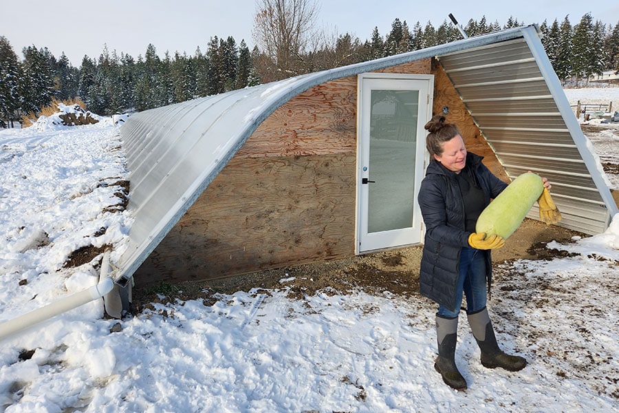 2023-27-greenhouse-in-the-snow-canada Winter view of geothermal greenhouse in Armstrong, BC with gardener holding large harvested squash outside entrance